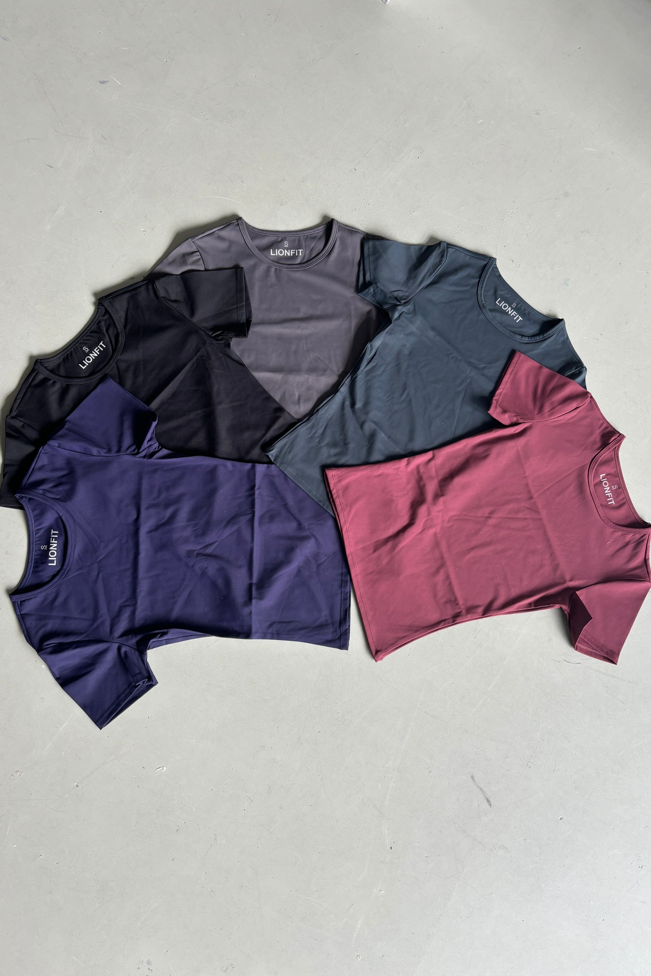 Essential Shortsleeve Top voor dames van Lion Fit Gymwear, stapel van vijf T-shirts in verschillende kleuren: donkerblauw, paars, zwart, grijs en rood. Perfect voor sporten en dagelijks gebruik.