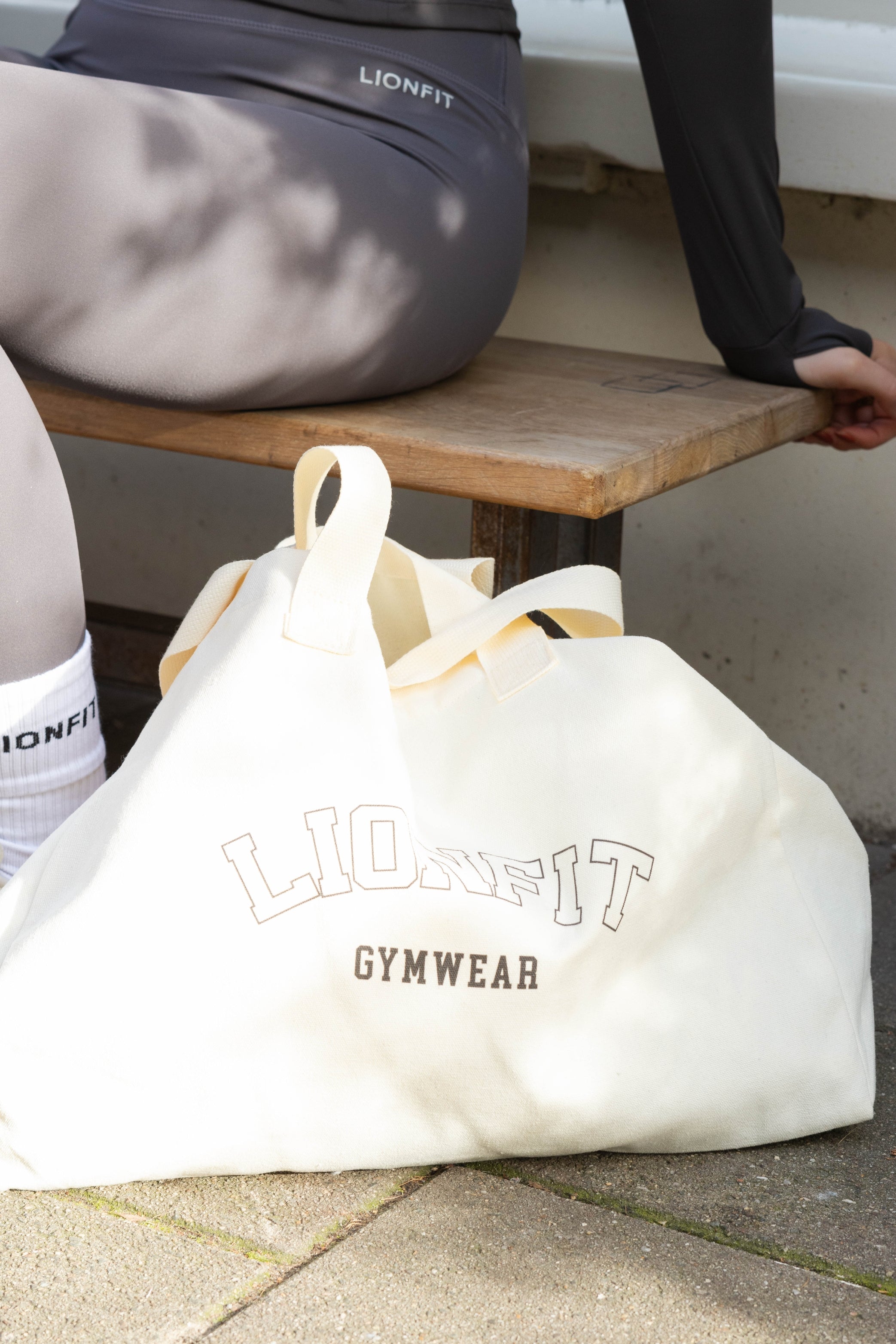 Vrouw met de Essential Totebag Beige van Lionfit Gymwear in sportieve outfit, buiten bij daglicht. Grote katoenen shopper tas perfect voor sportschool, strand of dagelijks gebruik, gemaakt van duurzaam gerecycled materiaal.