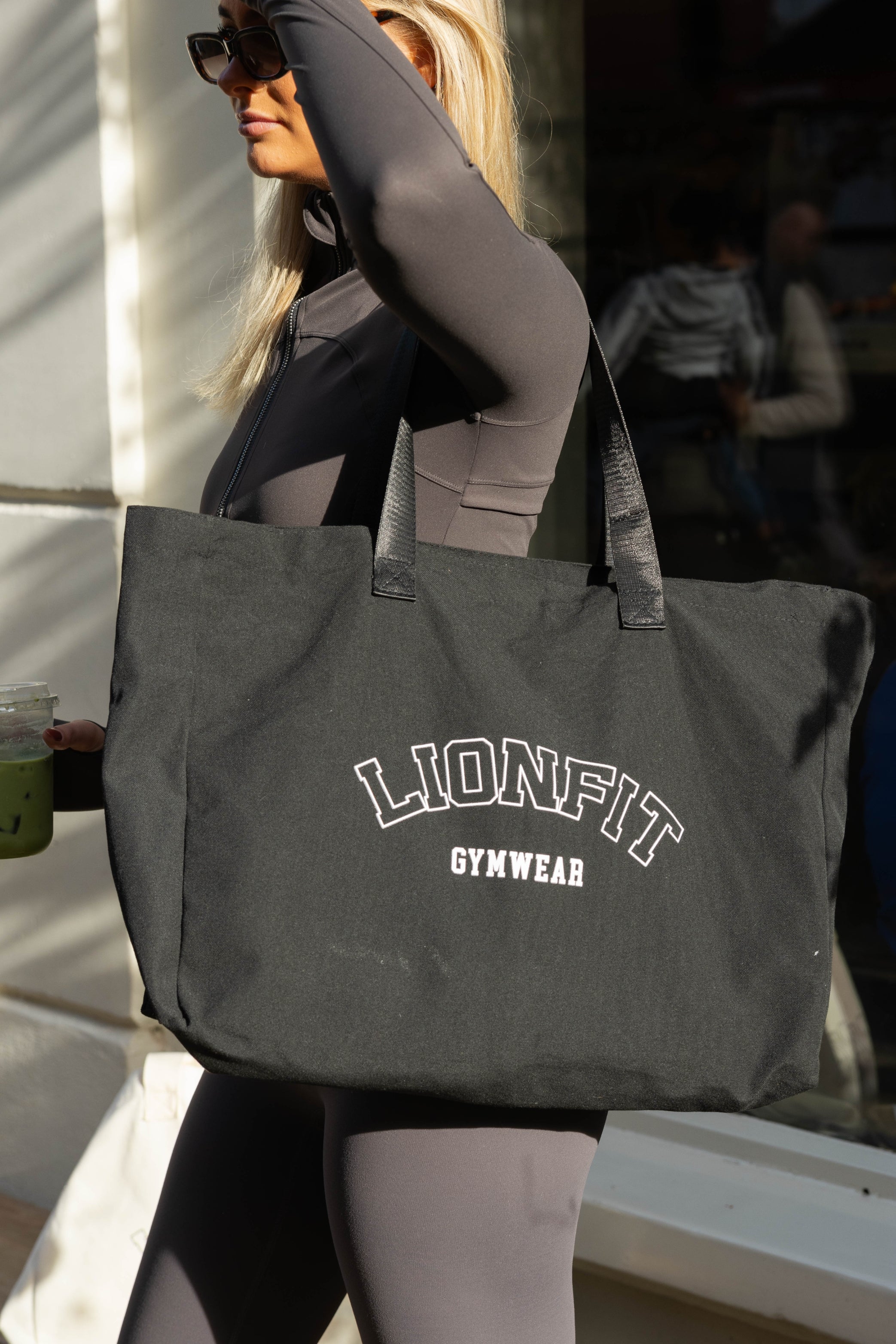 Vrouw met de zwarte Essential Totebag van Lionfit Gymwear, een grote katoenen shopper ideaal voor sportschool, dagelijks gebruik en strand. Gemaakt van gerecycled materiaal, biedt deze duurzame tas een stijlvolle en praktische oplossing voor actieve vrouwen.