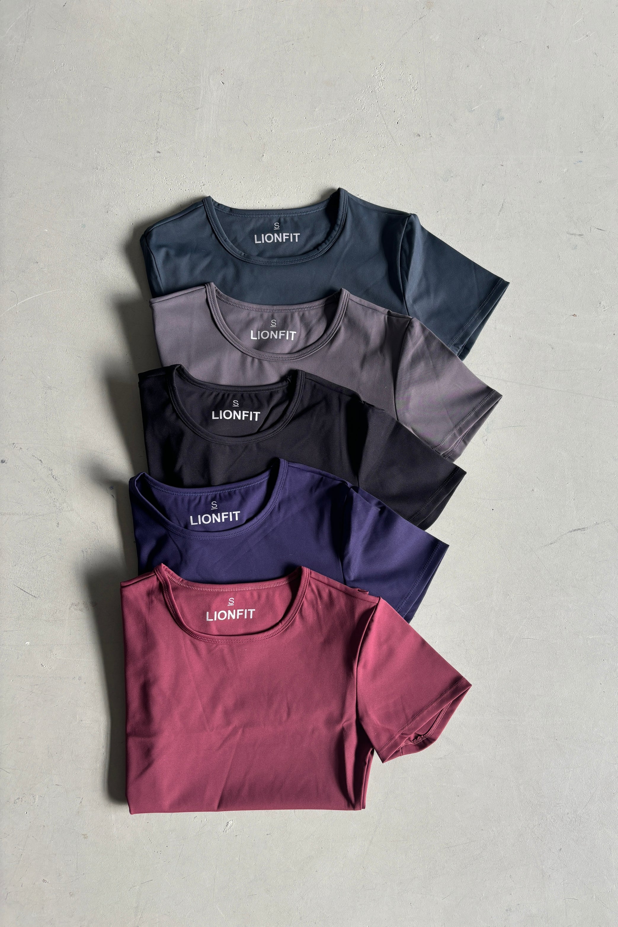 Essential Shortsleeve Top voor dames van Lion Fit Gymwear, stapel van vijf T-shirts in verschillende kleuren: donkerblauw, paars, zwart, grijs en rood. Perfect voor sporten en dagelijks gebruik.